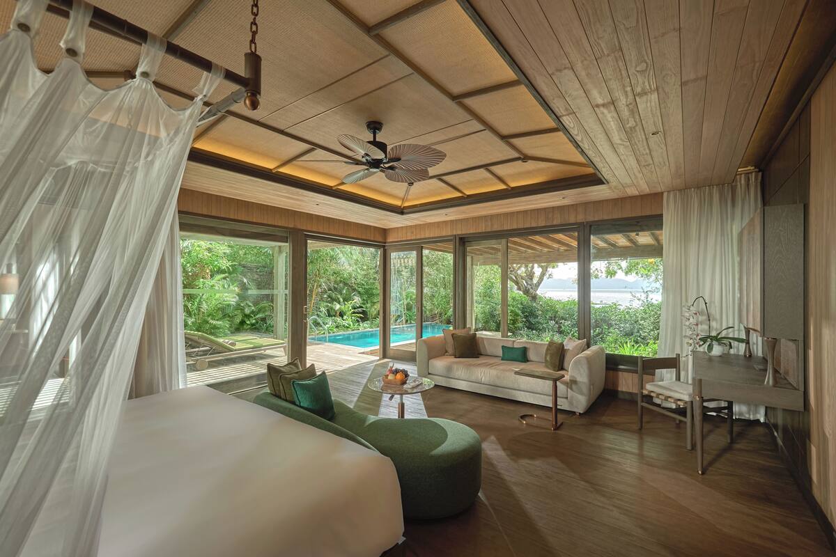 Ein geräumiges Schlafzimmer mit einem großen Bett, einem grünen Hocker, einem kleinen Tisch und Blick auf einen Swimmingpool und üppiges Grün.