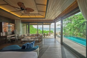 A spacious living area with a large sofa, a coffee table, and a TV. There is a wooden ceiling fan, a desk with a chair, and a view of a swimming pool and greenery outside. - An Lam Retreats Ninh Van Bay (Ninh Hoa)