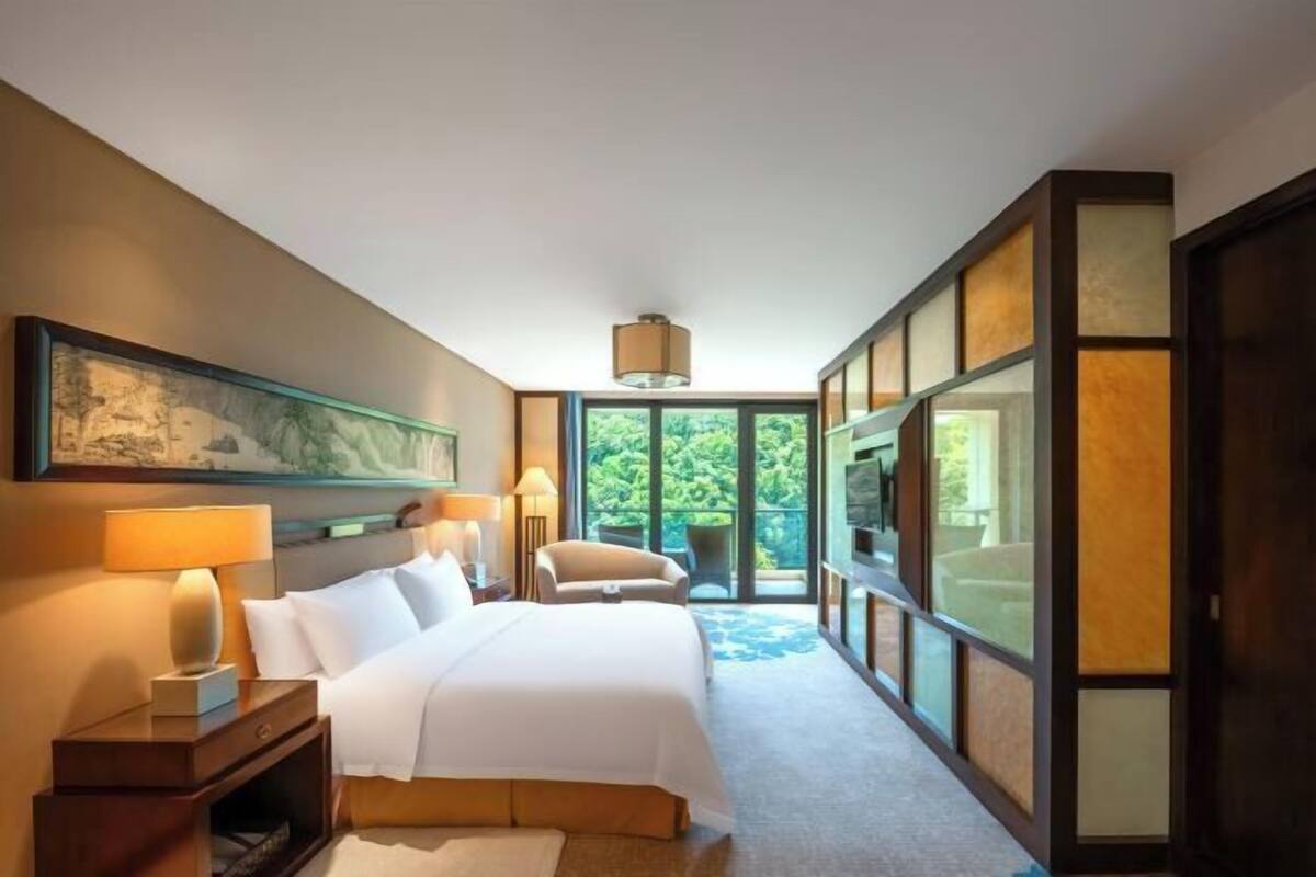 Een hotelkamer met een groot bed, nachtlampjes, een stoel, een groot raam en een schilderij aan de muur.
