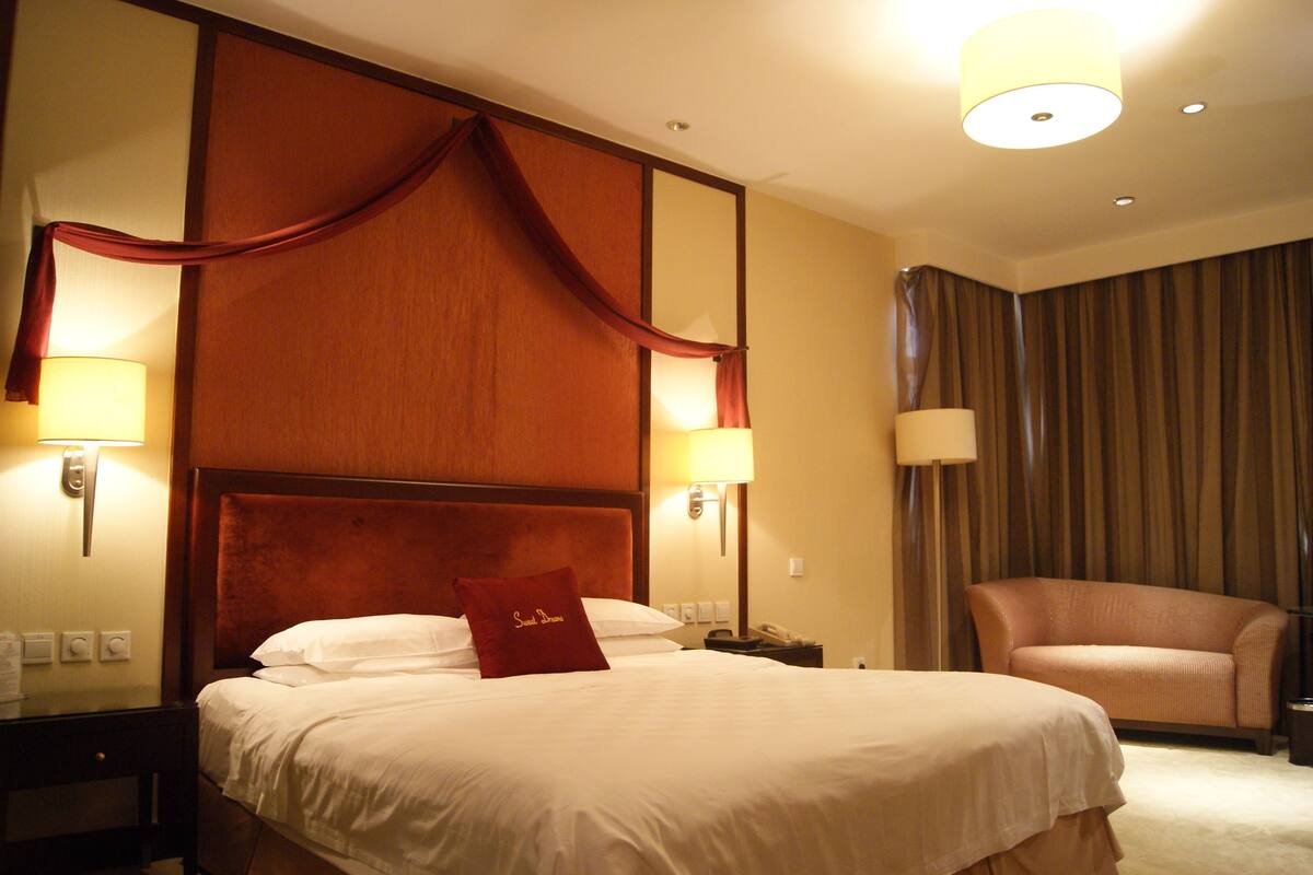 Een hotelkamer met een groot bed, twee bedlampen, een nachtkastje, een bank en een gordijn.