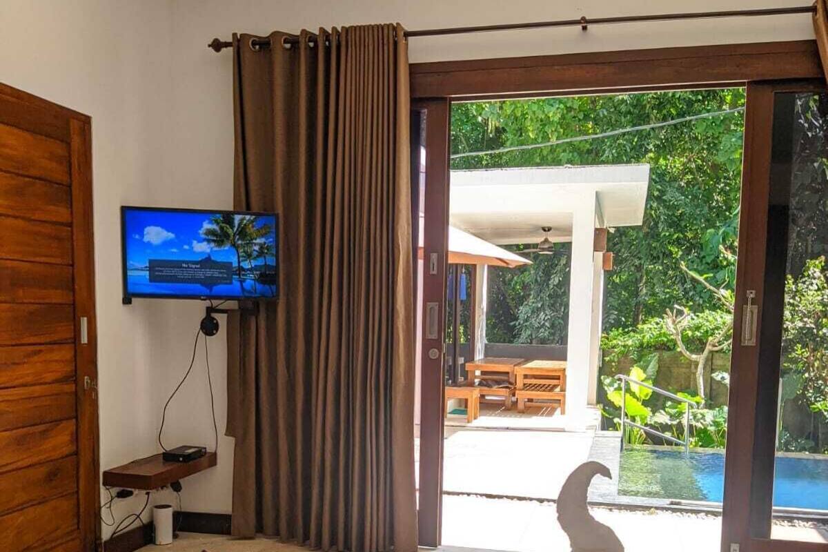 Habitación de hotel con cama, televisión en la pared, cortina y vista a una piscina y jardín a través de puertas corredizas de vidrio.