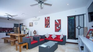 A modern living room with a sofa, coffee table, and wooden bench. There is a kitchen area with cabinets and a refrigerator. - Villa Collard (Senggigi)