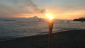 A beach scene with a torch in the foreground, a sunset in the background, and boats on the water. - Villa Collard (Senggigi)