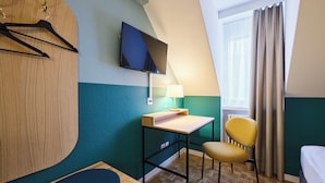 Habitación de hotel con cama, escritorio con lámpara, una silla, televisor montado en la pared y ventana con cortinas.