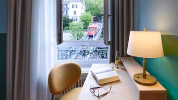 Una habitación con un escritorio de madera, una silla, un ventanal con vistas a un tren rojo y edificios, y una lámpara.