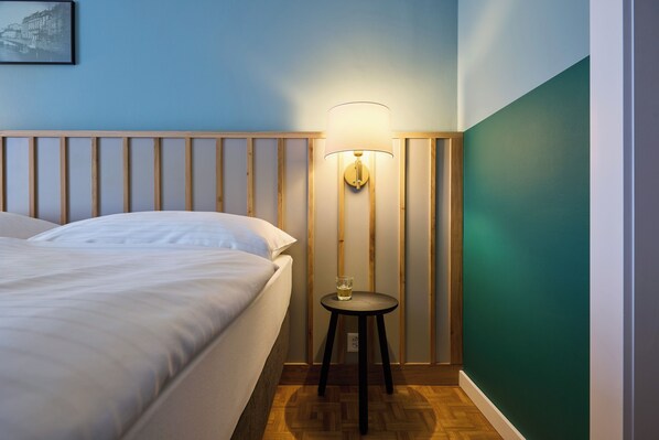 Una cama bien hecha con ropa blanca, una mesita redonda con una lámpara y un cuadro colgado en la pared.