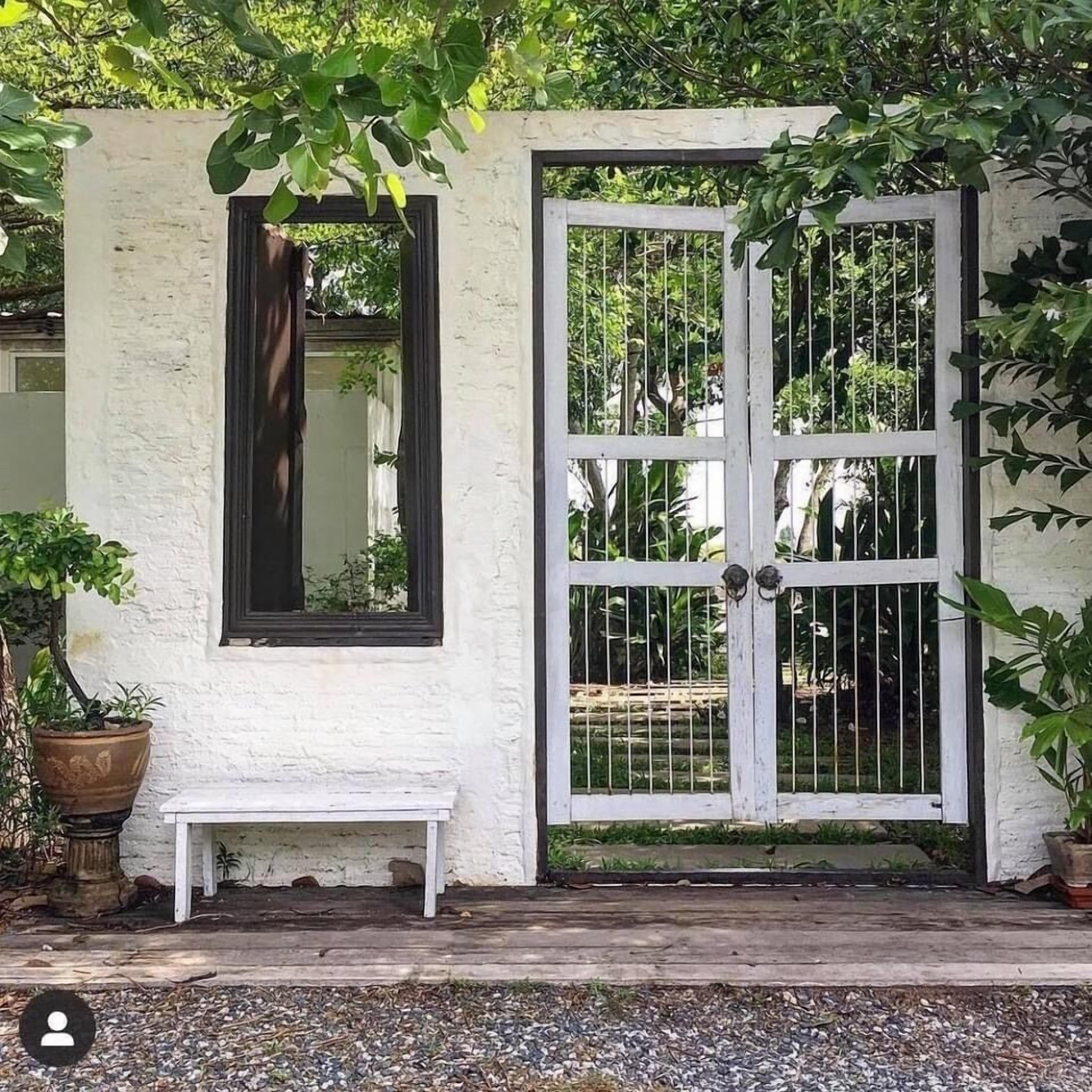Un edificio blanco con un portón metálico grande, una banca y plantas en macetas.