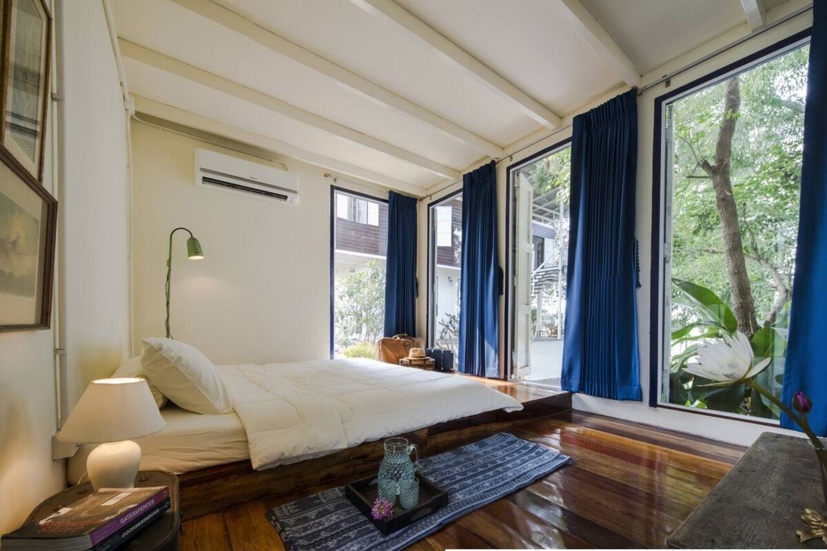 A bedroom with a bed, a nightstand, a lamp, a window with curtains, and a view of greenery outside.