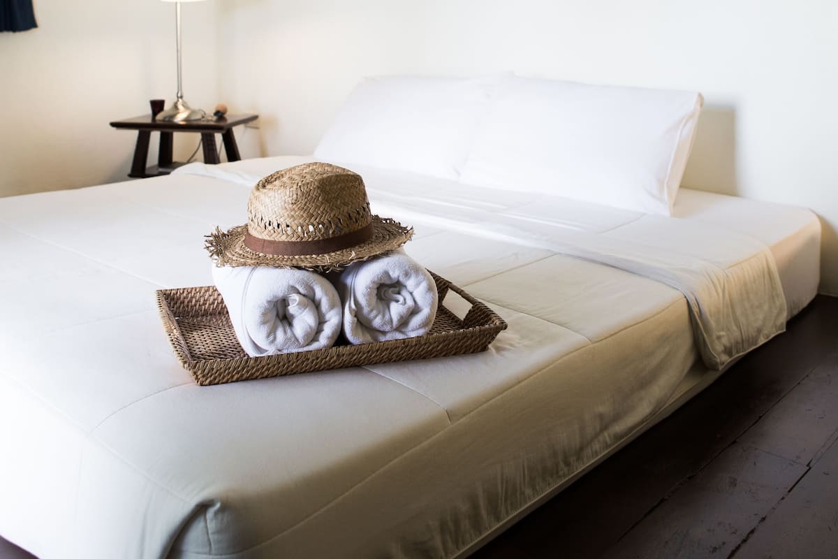 A neatly made bed with a straw hat and towels on a tray, a lamp on a nightstand, and a wall with framed pictures.