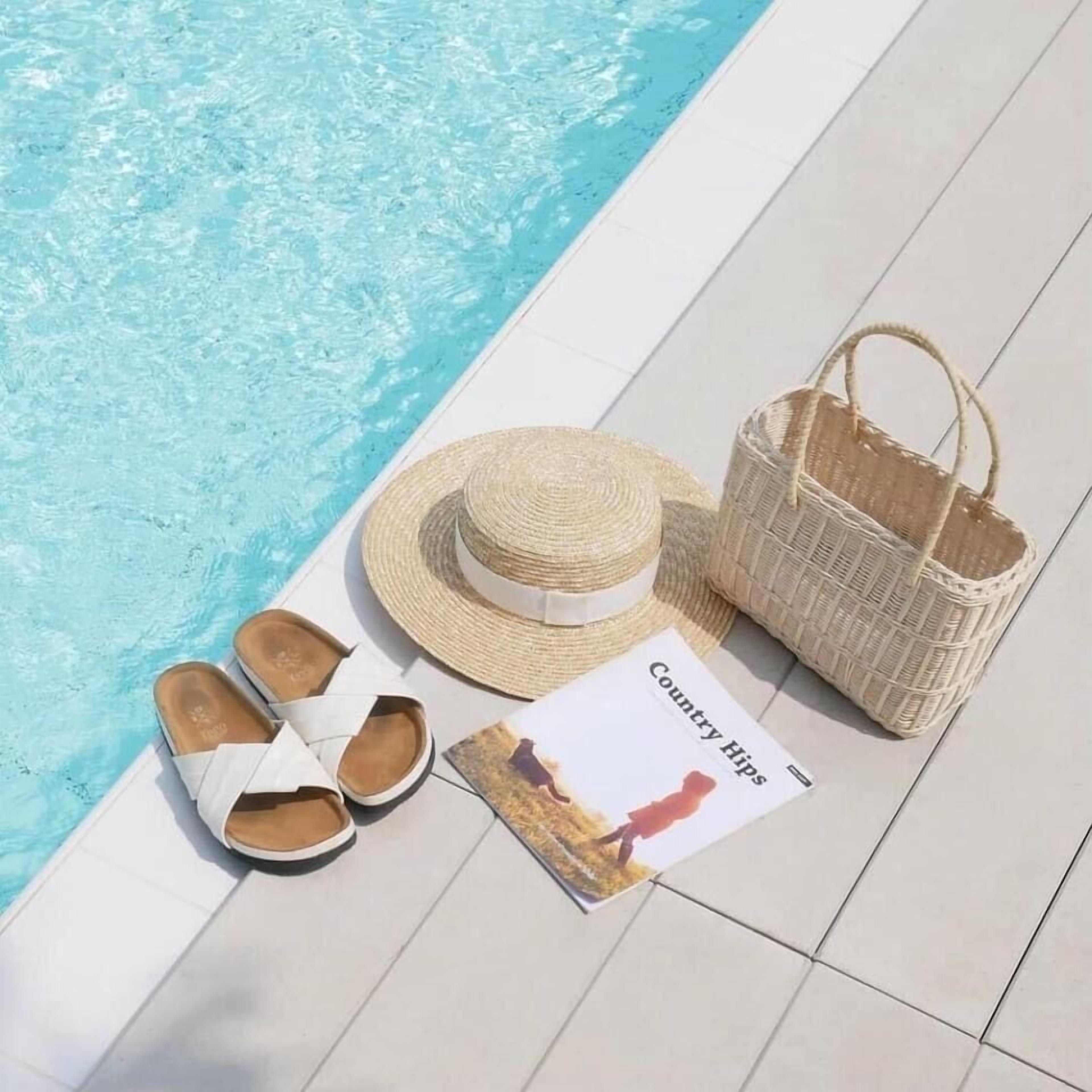 Escena junto a la piscina con un sombrero de paja, un bolso tejido, sandalias blancas y una revista titulada "Country Hips".