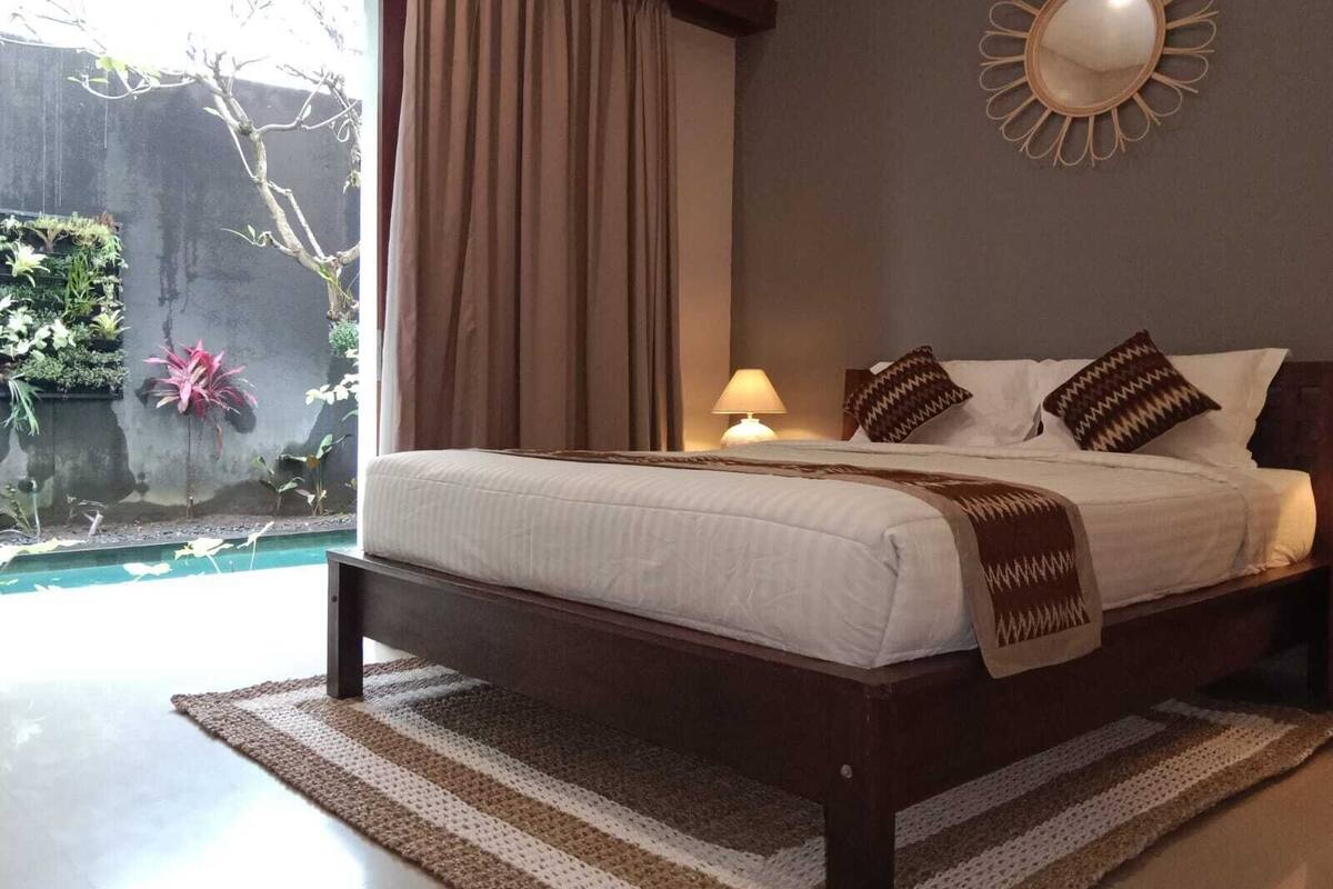 A bedroom with a bed, curtains, a nightstand, a lamp, a mirror, and a view of a garden and pool.