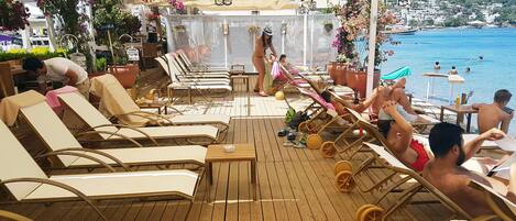 A wooden deck with sun loungers and tables, overlooking a body of water with people swimming.