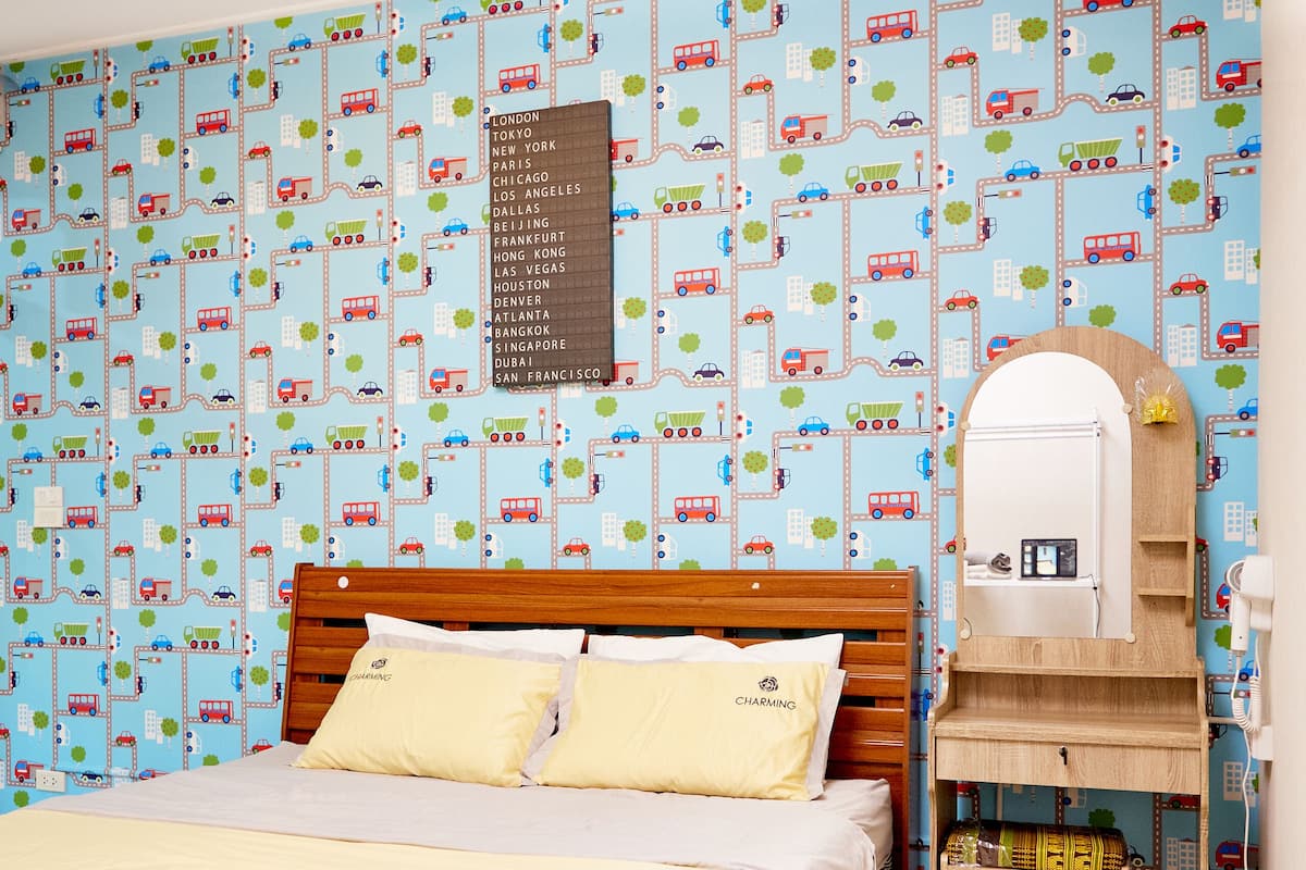 A neatly made bed with a wooden headboard, a bedside table with a mirror, and a wall with a travel-themed wallpaper.