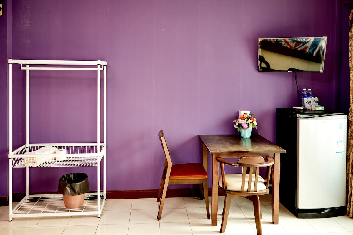A room with a purple wall, a white metal shelf unit, a wooden dining table with two chairs, a small refrigerator, and a TV mounted on the wall.