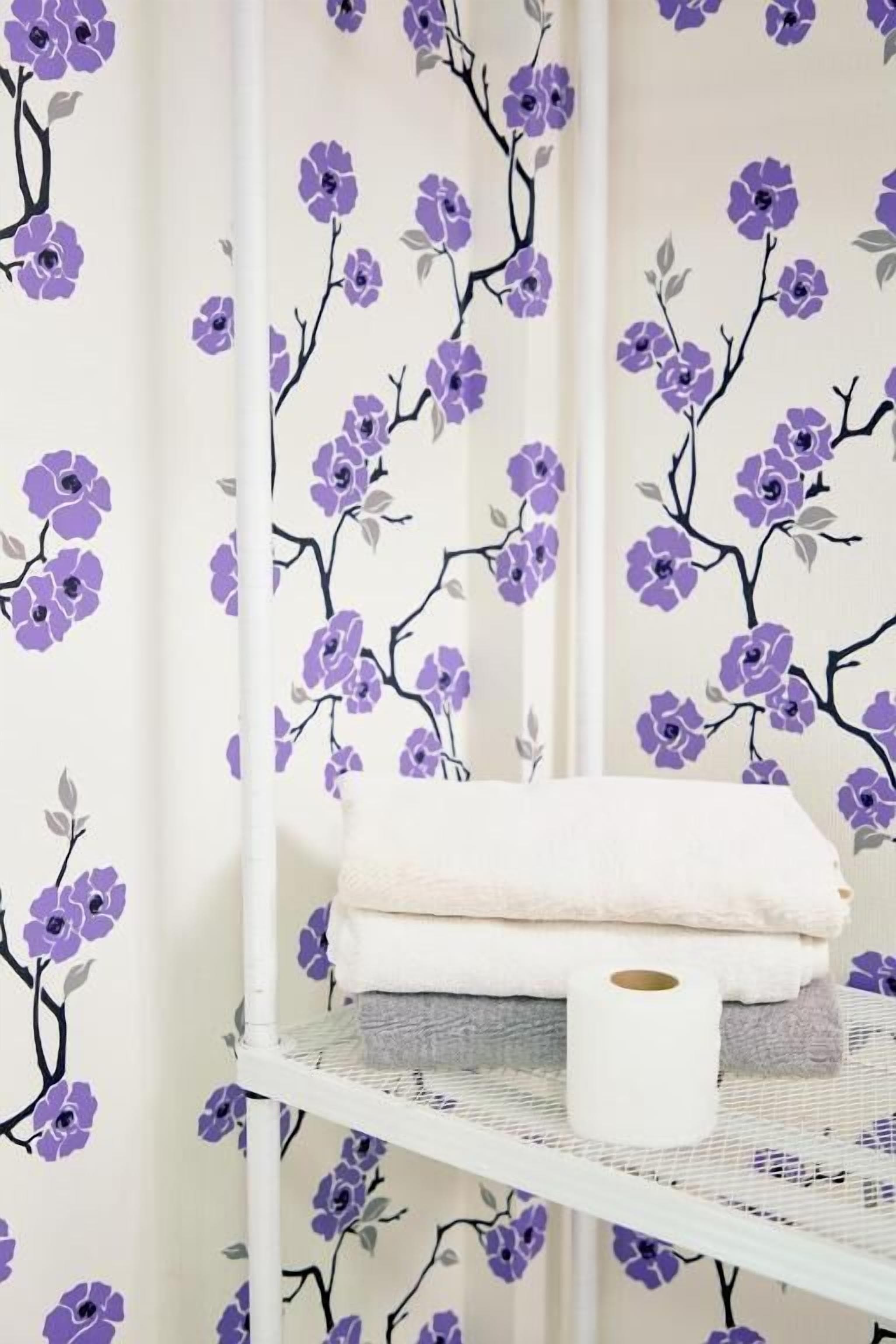 A bathroom with a white shower curtain featuring a purple floral pattern, a white towel, and a roll of toilet paper on a metal shelf.