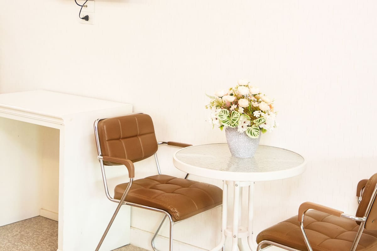 A room with a white desk, a round table with two brown chairs, a flower vase, and a mounted TV.