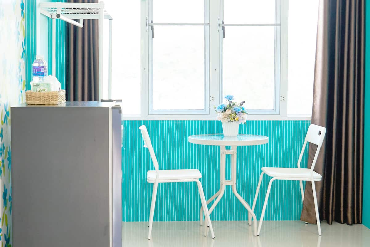 A room with a window, a table with two chairs, a refrigerator, and curtains.