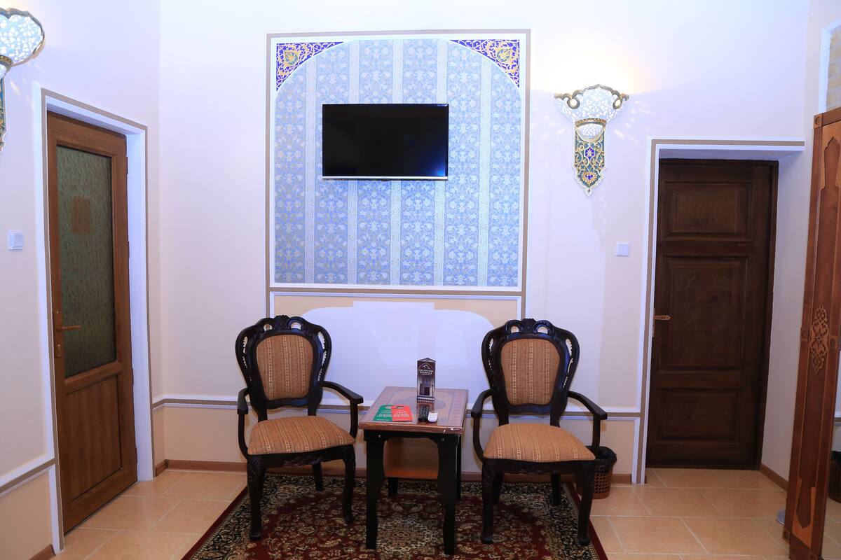 A room with two wooden chairs, a small table, a flat-screen TV mounted on the wall, and a patterned window.