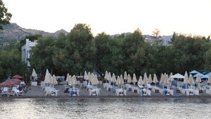 Een strandgebied met rijen ligbedden en parasols, een gebouw op de achtergrond en een waterpartij op de voorgrond.