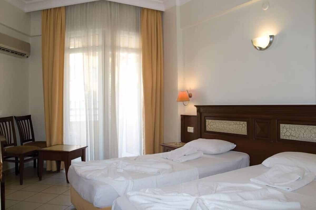 A hotel room with two beds, a desk, chairs, a window with curtains, and a wall-mounted lamp.