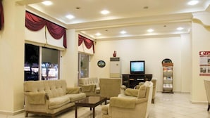 A living room with beige sofas, a coffee table, and a TV. There is a window with curtains, a vase on the wall, and a vending machine. - Alin Hotel (Alanya)