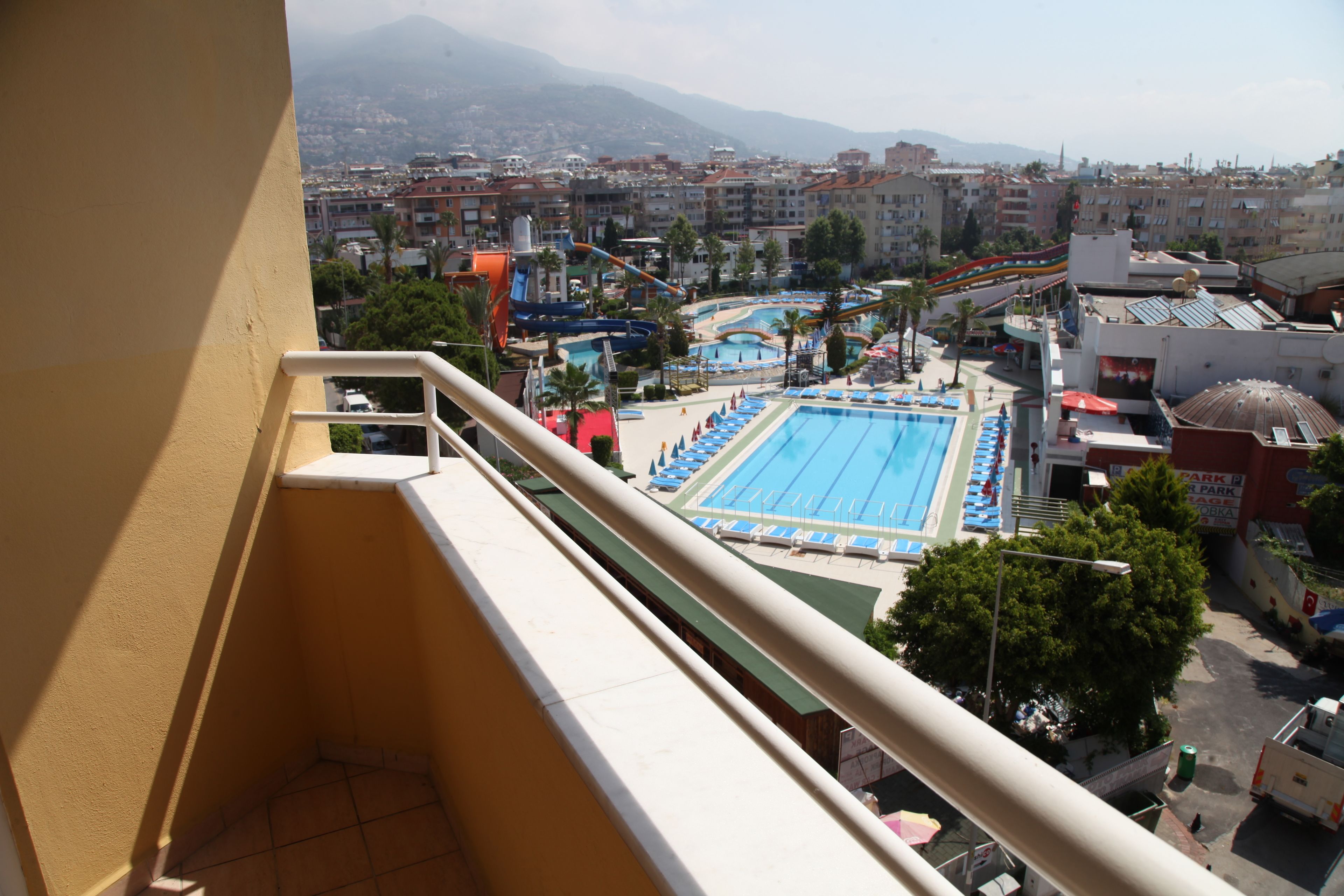 Vue depuis un balcon donnant sur un espace piscine avec un toboggan aquatique, entouré de bâtiments et avec une montagne en arrière-plan.