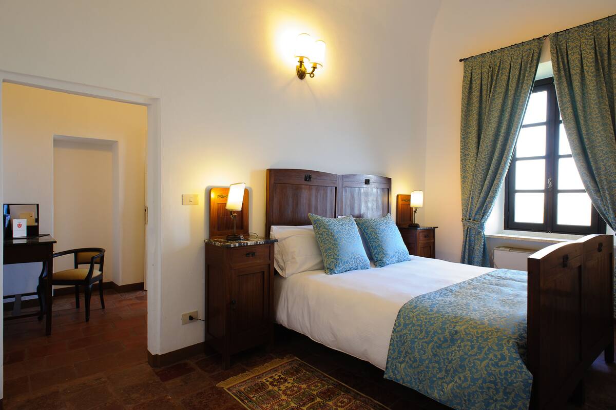 A bedroom with a wooden bed, two bedside lamps, a desk with a chair, and a window with curtains.