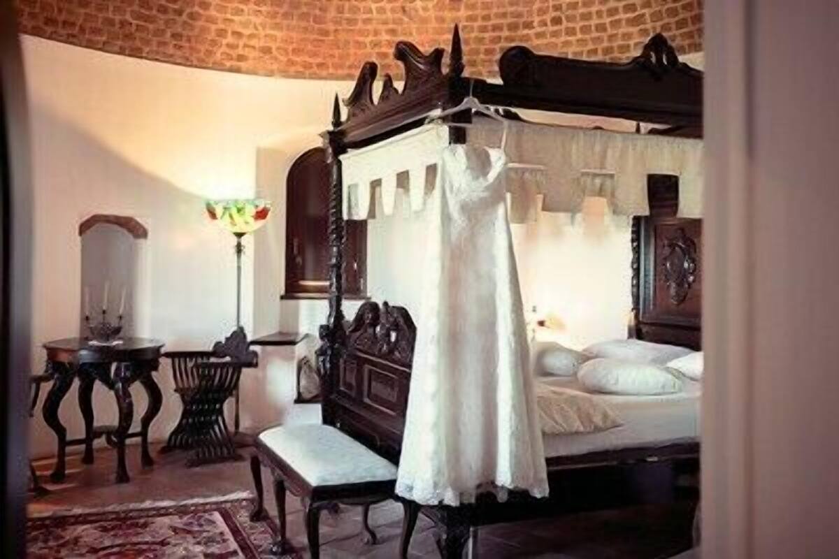 A traditional four-poster bed with a canopy, a small table with a lamp, and a chair in a room with brick walls and a tiled floor.