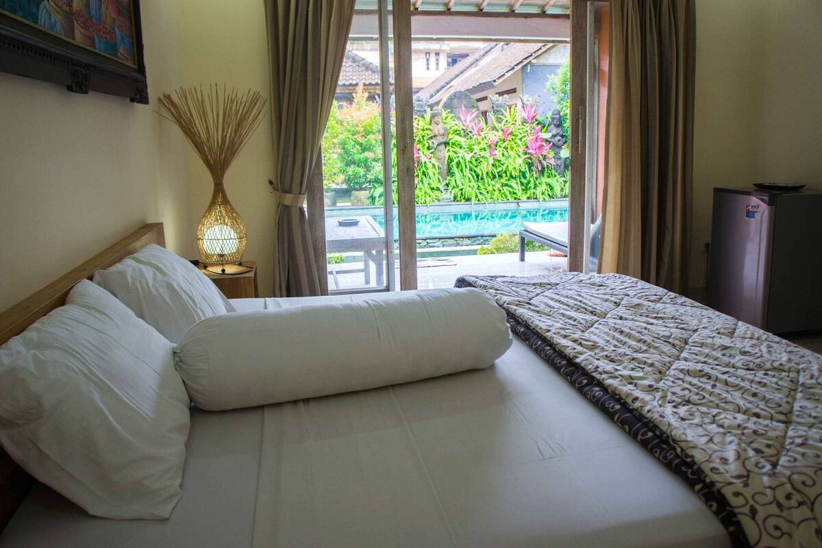A hotel room with a bed, a nightstand, a lamp, and a view of a garden through a sliding door.