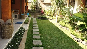 A courtyard with a stone pathway leading to a door, surrounded by greenery and a traditional building with a tiled roof. - Bale Bali Inn (Ubud)