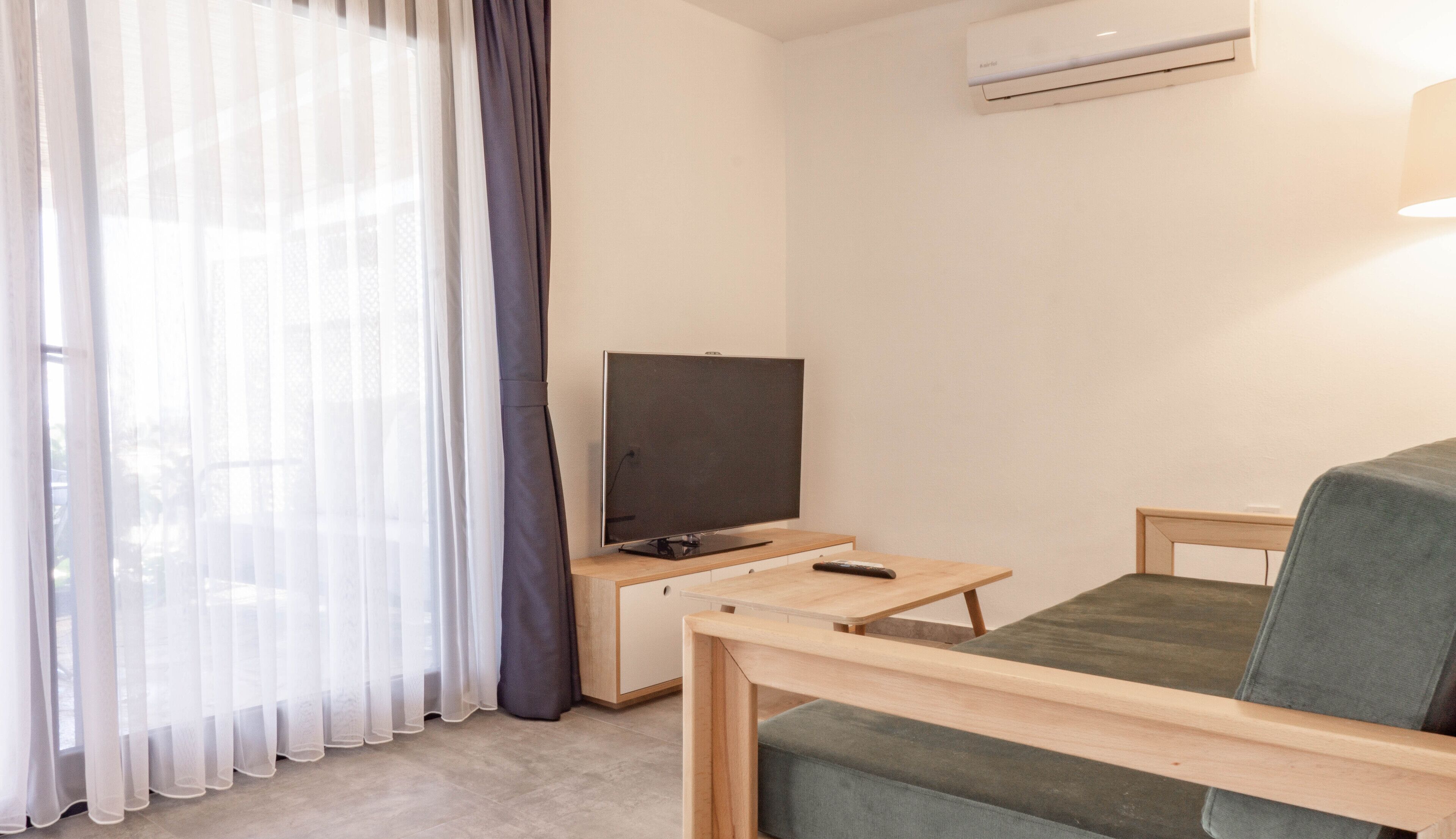 Una habitación moderna y minimalista con televisión de pantalla plana sobre un soporte de madera, una pequeña mesa de madera, un sofá verde y cortinas translúcidas.