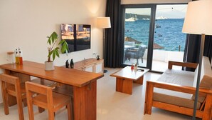 Una habitación de hotel moderna con una mesa de comedor de madera, dos sillas, un pequeño sofá y vistas al mar a través de amplias ventanas.