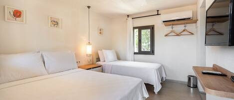 Une chambre d’hôtel avec deux lits, une table de chevet avec une lampe, une petite fenêtre avec des rideaux, un téléviseur fixé au mur et un banc en bois.