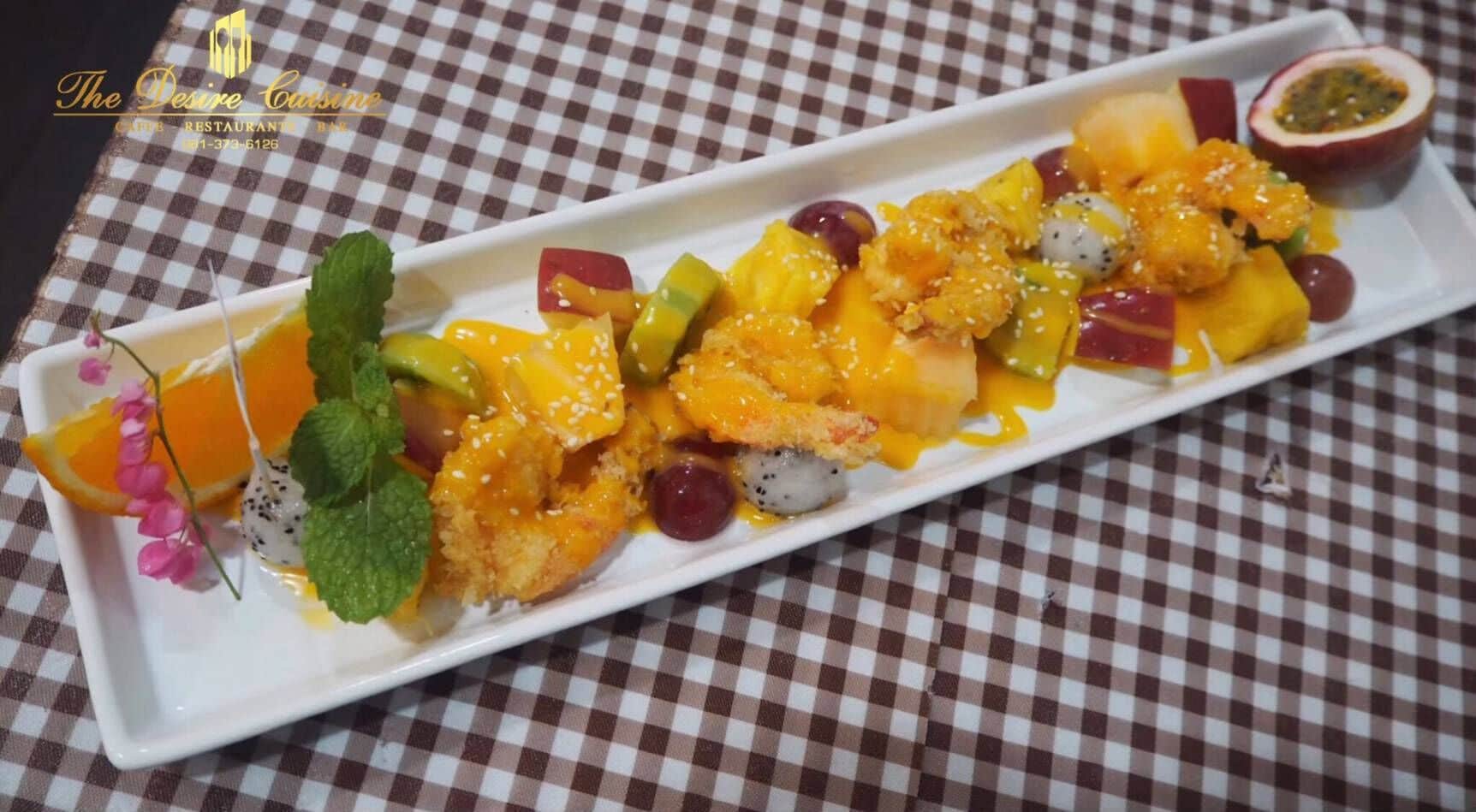 A white rectangular plate with a variety of fruits and a dessert, garnished with mint leaves and a small flower, on a checkered tablecloth.