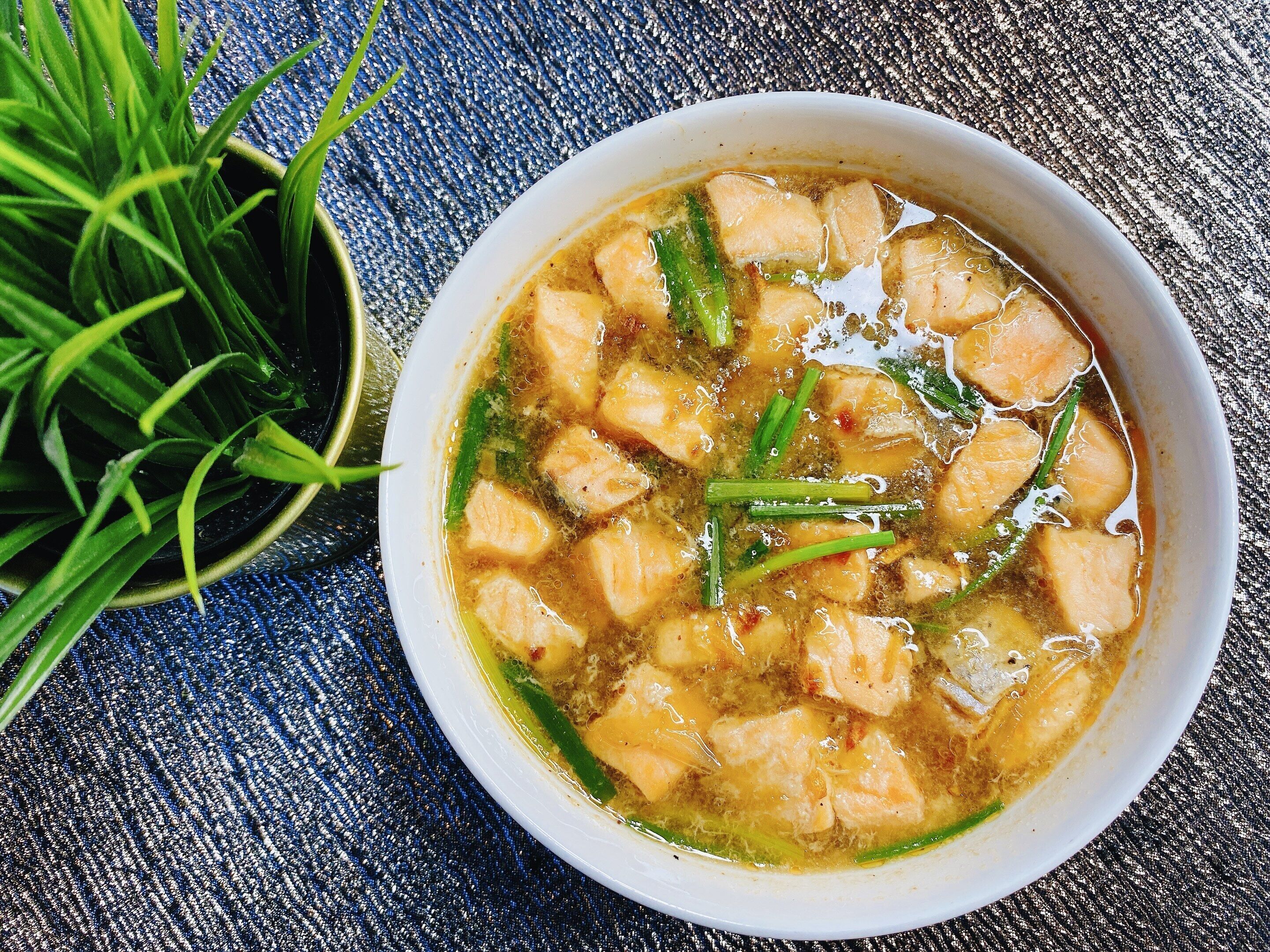 Un bol de soupe contenant du tofu et des oignons verts, posé sur une surface bleue texturée.