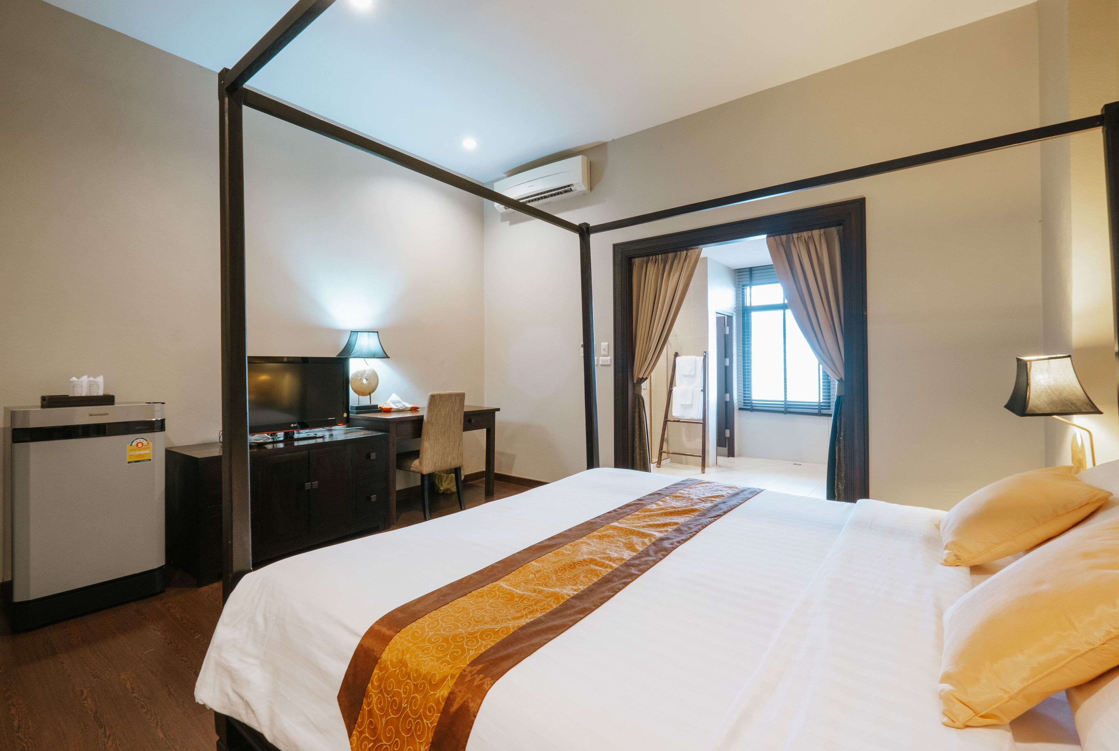 A hotel room with a large bed, a desk with a chair, a small refrigerator, and an air conditioning unit.