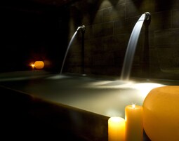 Zona de spa tenuemente iluminada con una bañera, dos chorros de agua y tres velas encendidas.