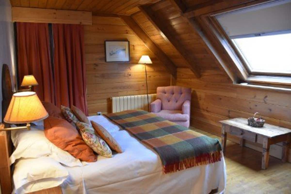 Ein gemütliches Schlafzimmer im Dachgeschoss mit schrägen Wänden, einem Bett mit bunter Decke, einem rosa Sessel und einem kleinen Tisch mit einem Korb.