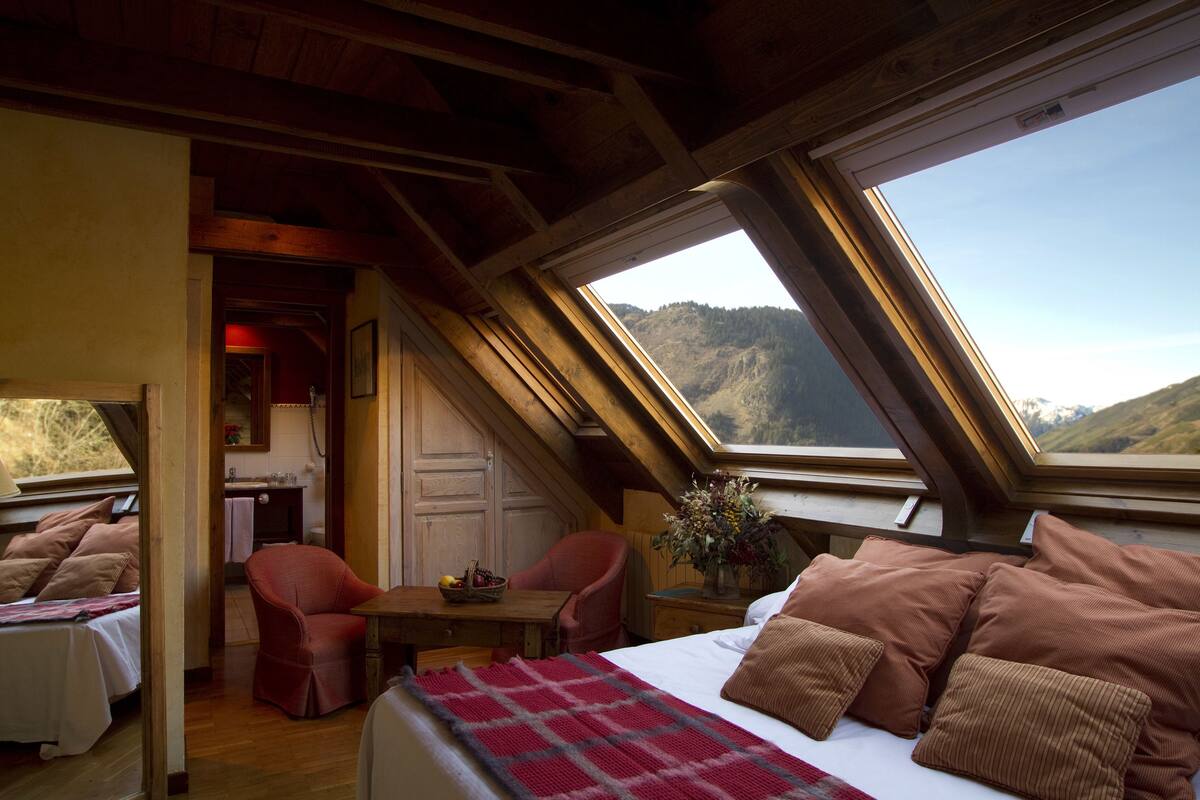Ein Schlafzimmer mit einem großen Bett, einem roten Sessel, einem hölzernen Schreibtisch und Blick auf eine gebirgige Landschaft durch Dachfenster.