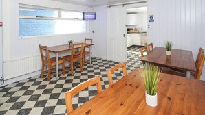 Une salle à manger avec un sol à carreaux, des tables et des chaises en bois. On y trouve une fenêtre donnant sur un mur de briques, une porte menant à la cuisine et un panneau indiquant l’endroit où se laver les mains.