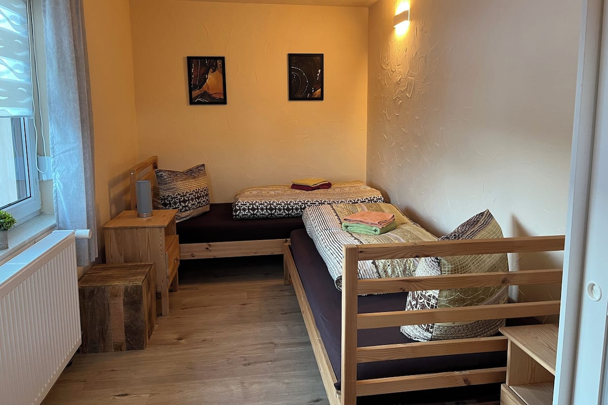 Um quarto pequeno e acolhedor com uma cama de solteiro, uma mesinha de cabeceira, uma janela com persianas e uma luminária fixa na parede.
