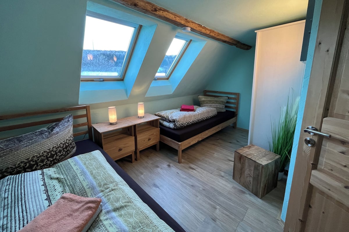 Quarto com teto inclinado, claraboias, cama com roupa de cama estampada, mesas de cabeceira com abajures e guarda-roupa de madeira.