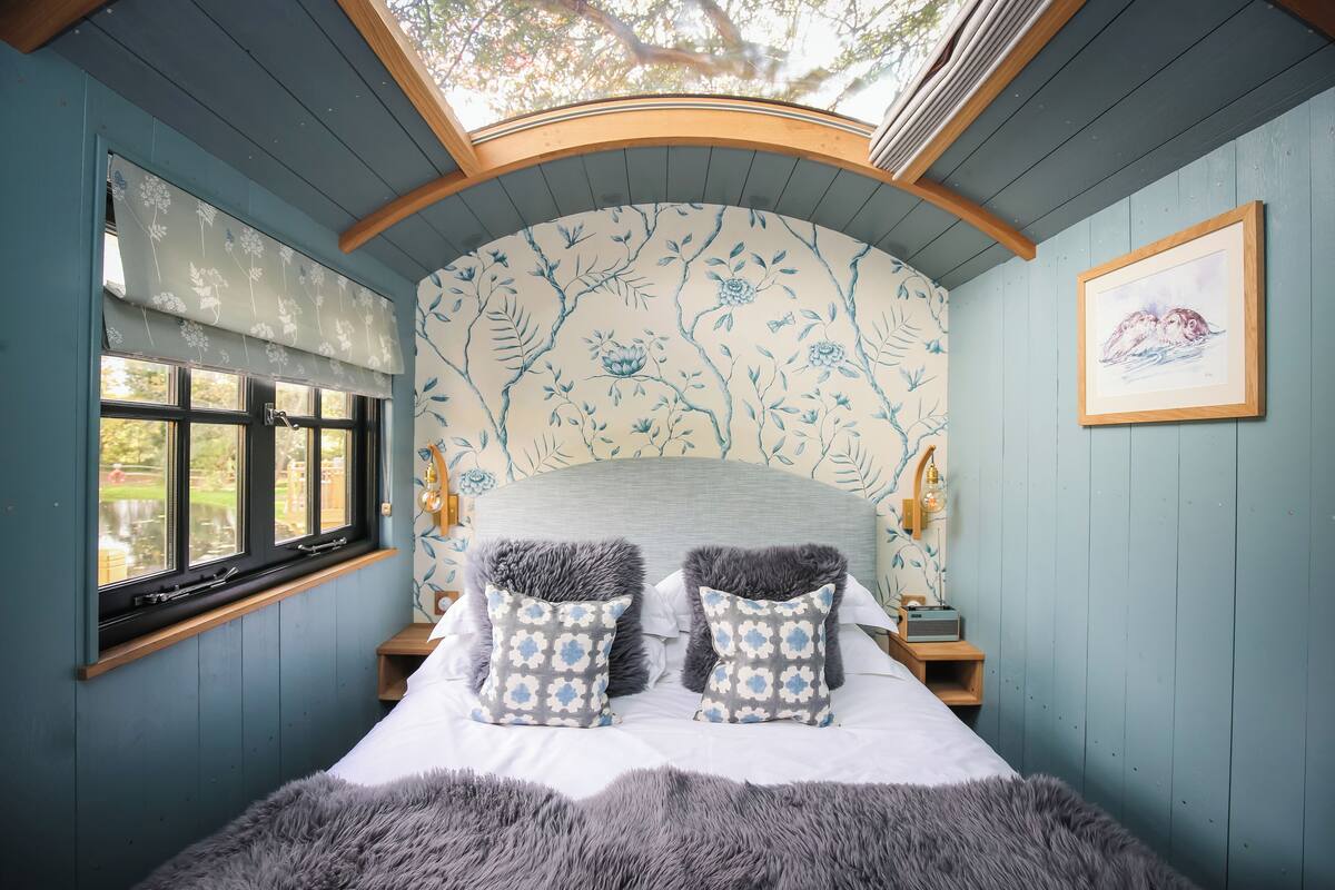 A bedroom with a skylight, a bed with patterned pillows, a wooden nightstand, and a framed picture on the wall.