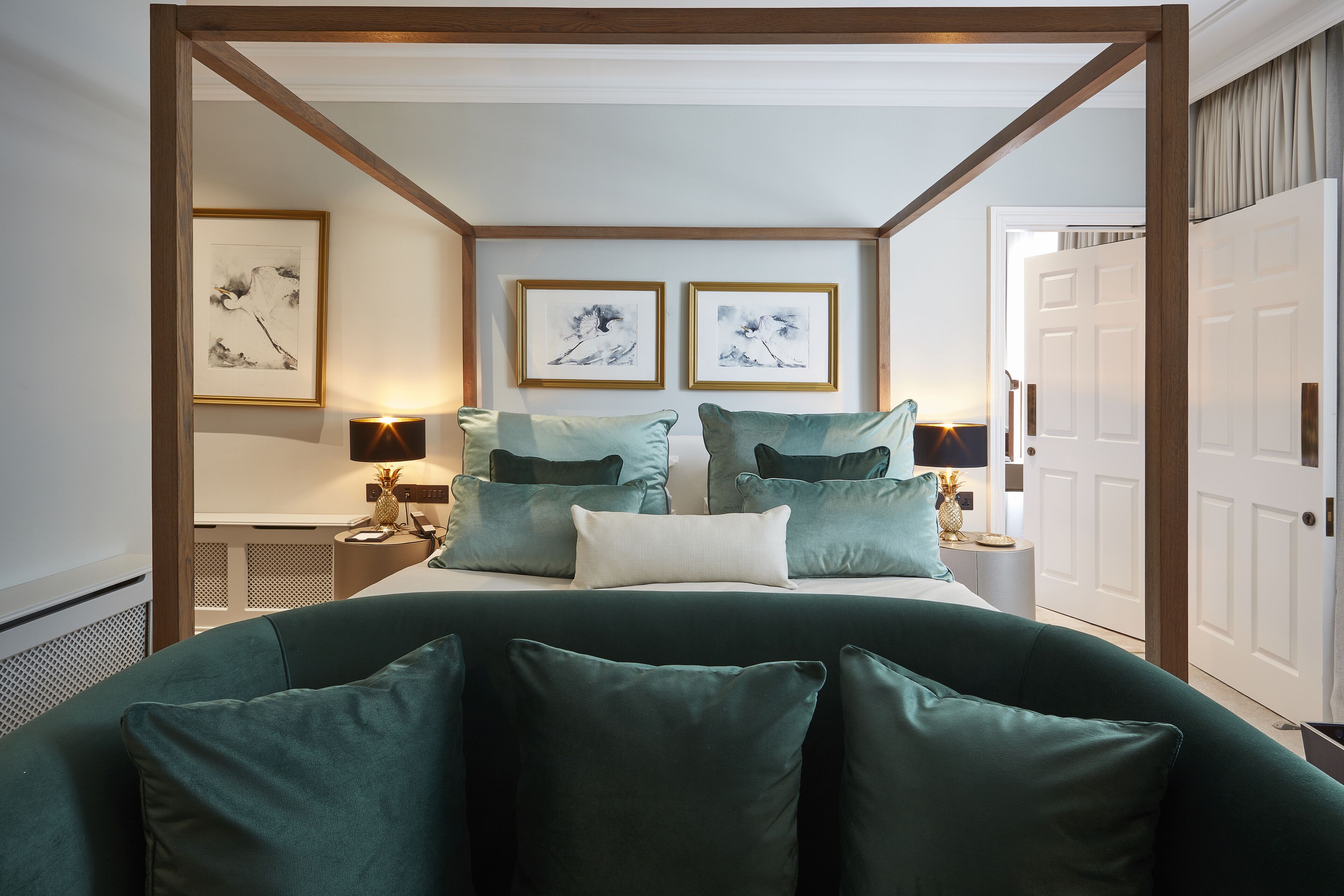 A bedroom with a four-poster bed, two framed artworks on the wall, and a nightstand with a lamp.