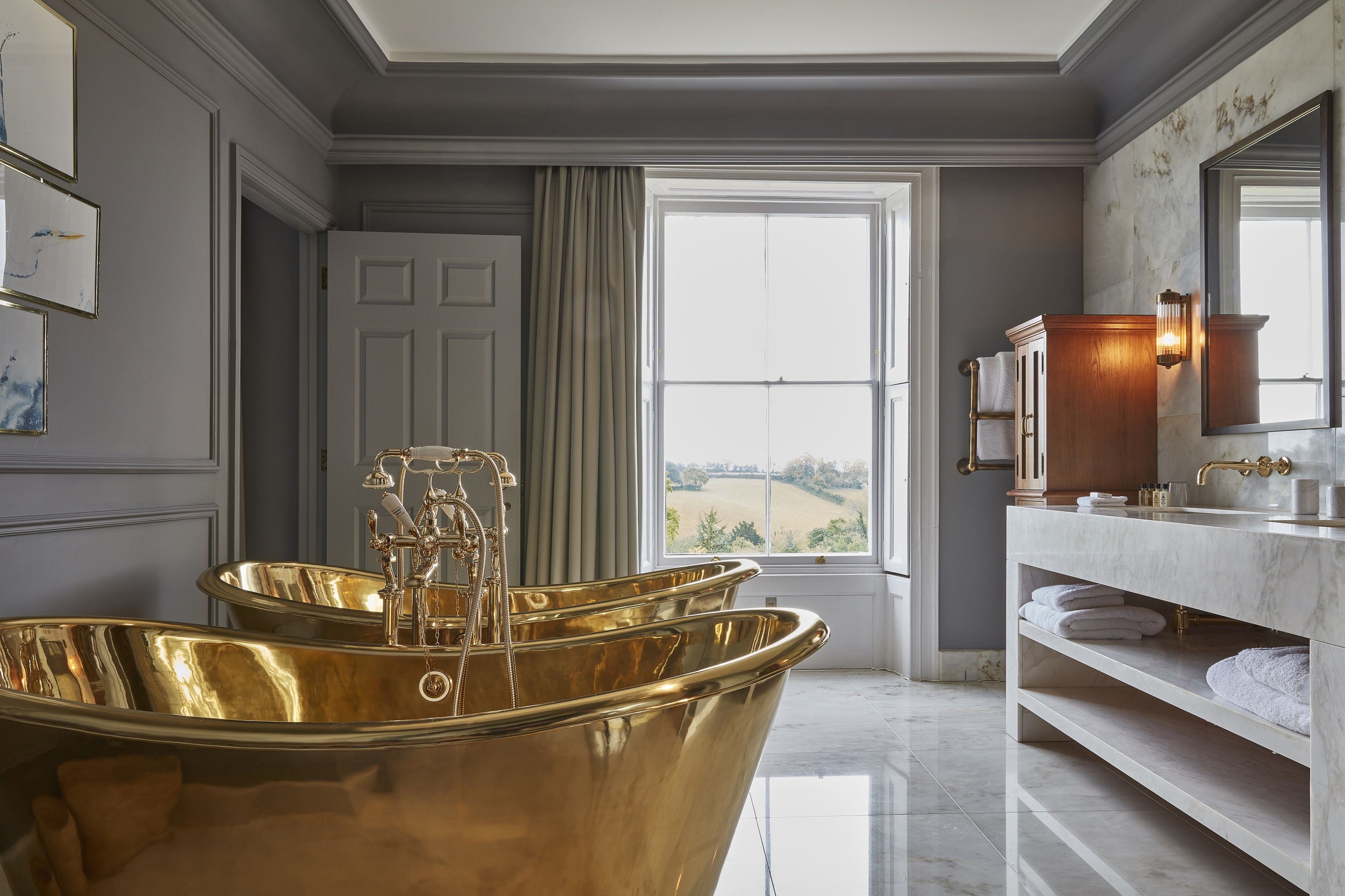 Une salle de bain luxueuse avec une grande baignoire dorée, un meuble-lavabo en marbre et une vue sur la campagne par une grande fenêtre.