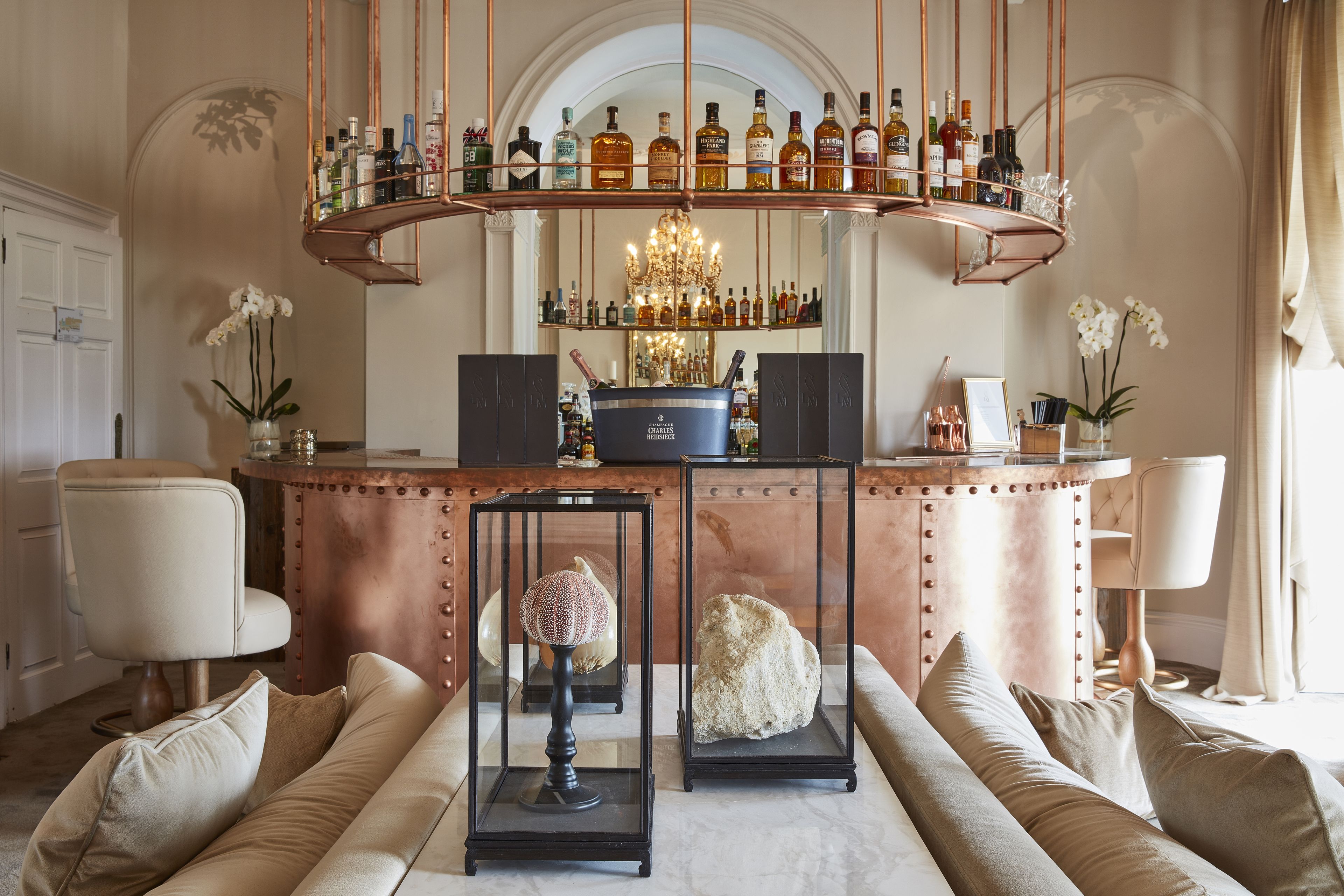 A bar area with a copper bar counter, a chandelier, and a variety of bottles. There are cushioned seating areas in front of the bar.