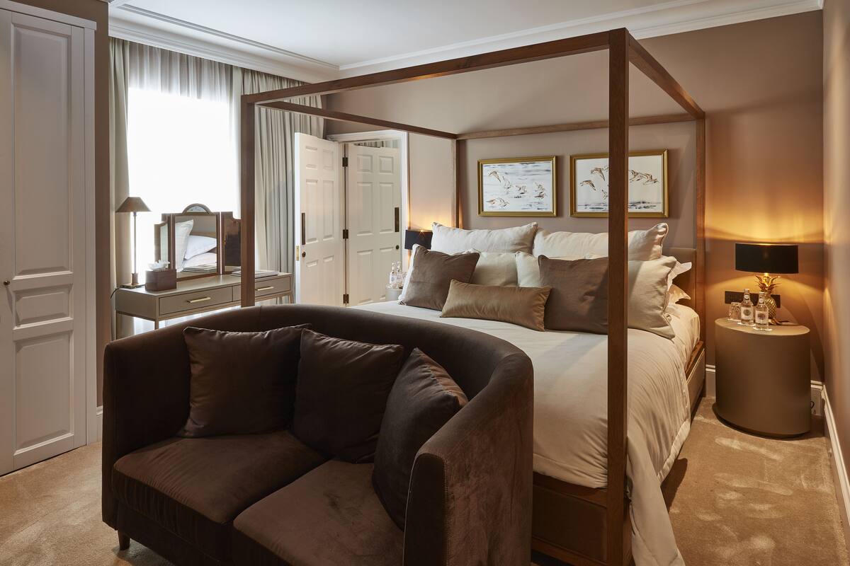 A four-poster bed with a headboard, a brown sofa, a bedside table with a lamp, and a framed artwork on the wall.