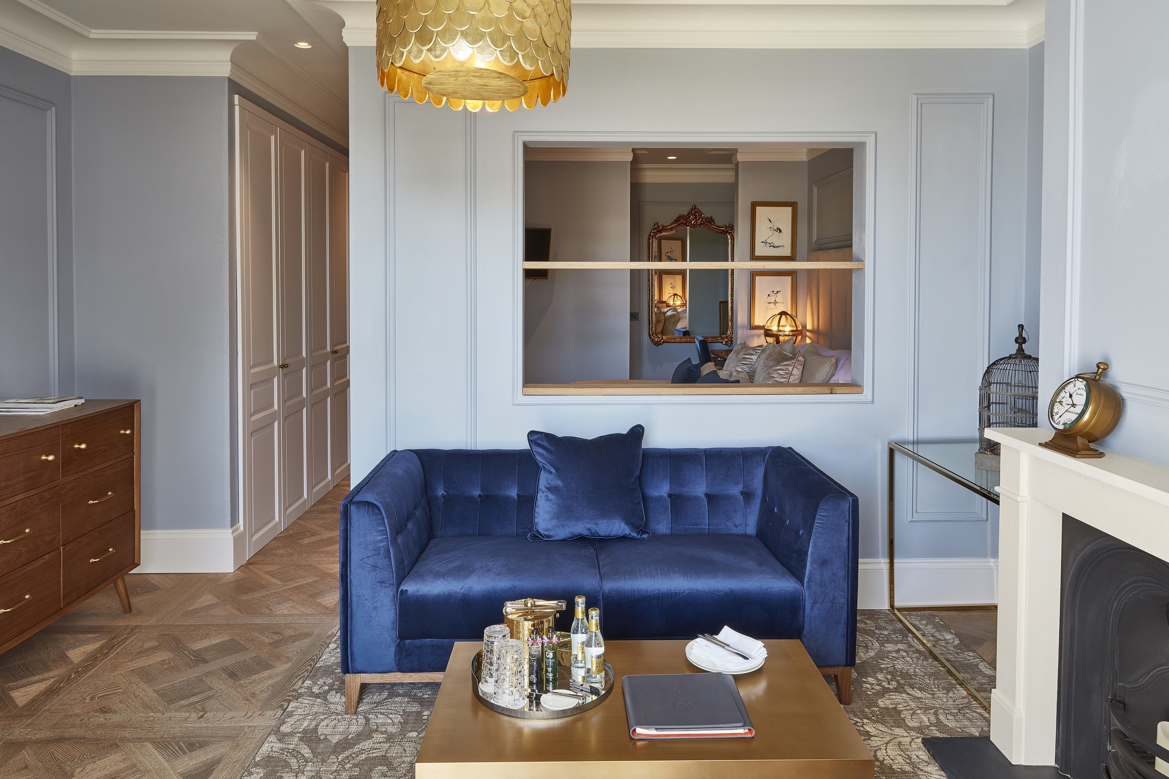 Un salon avec un canapé bleu, une table basse en bois sur laquelle sont posées des bouteilles, une cheminée et une grande fenêtre avec un miroir.