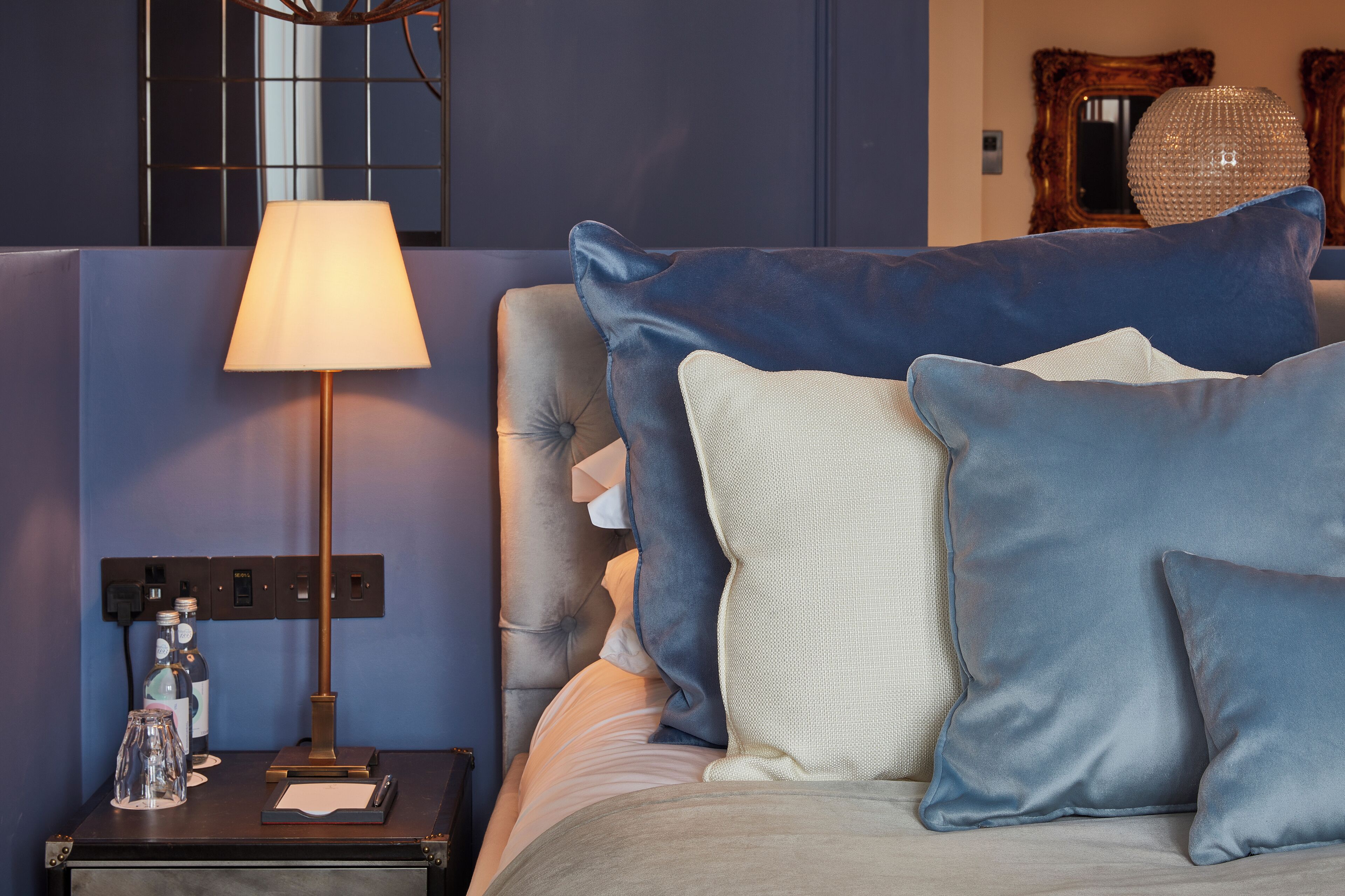 Een net opgemaakt bed met blauwe en witte kussens, een bedlampje en een klein tafeltje met flessen en een notitieboekje.