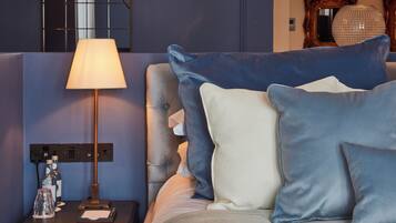 Een net opgemaakt bed met blauwe en witte kussens, een bedlampje en een klein tafeltje met flessen en een notitieboekje.